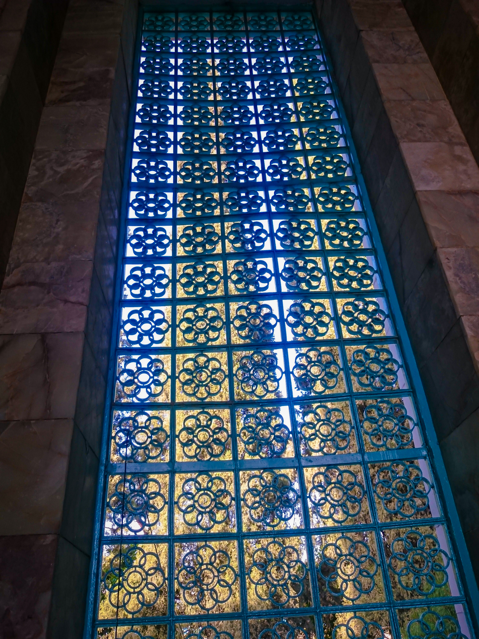 A close-up view of a frosted glass window with a blue square metal grid. Each square contains a stylized, hollow-petal floral design. The window is set in a white marble wall, and blurred greenery is visible through the glass.