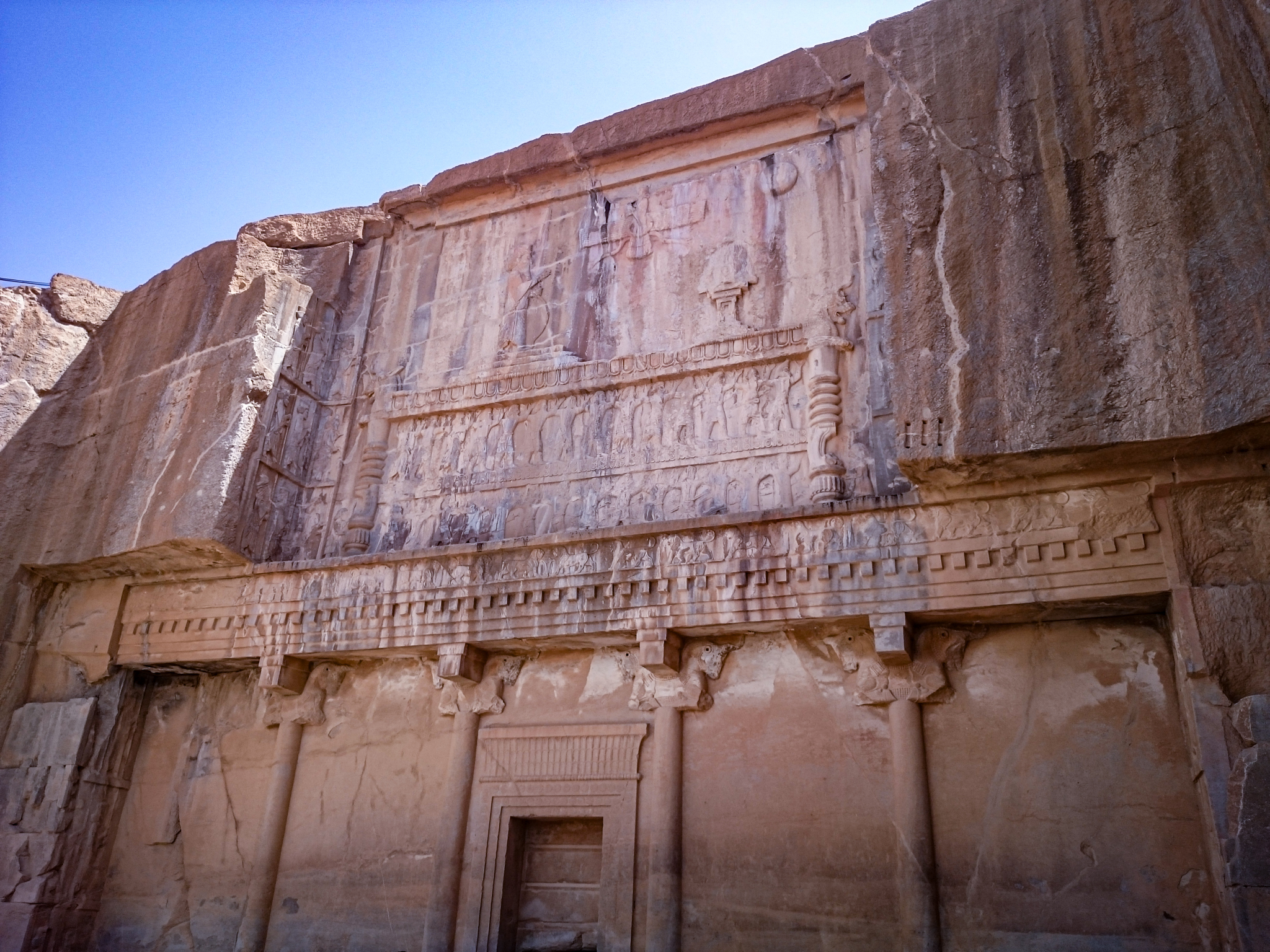 A monumental, cross-shaped tomb facade carved directly into a rocky cliffside at Persepolis. The entrance is visible in the center, with relief carvings above, resembling a palace structure. The stone is weathered, showing its ancient origins.