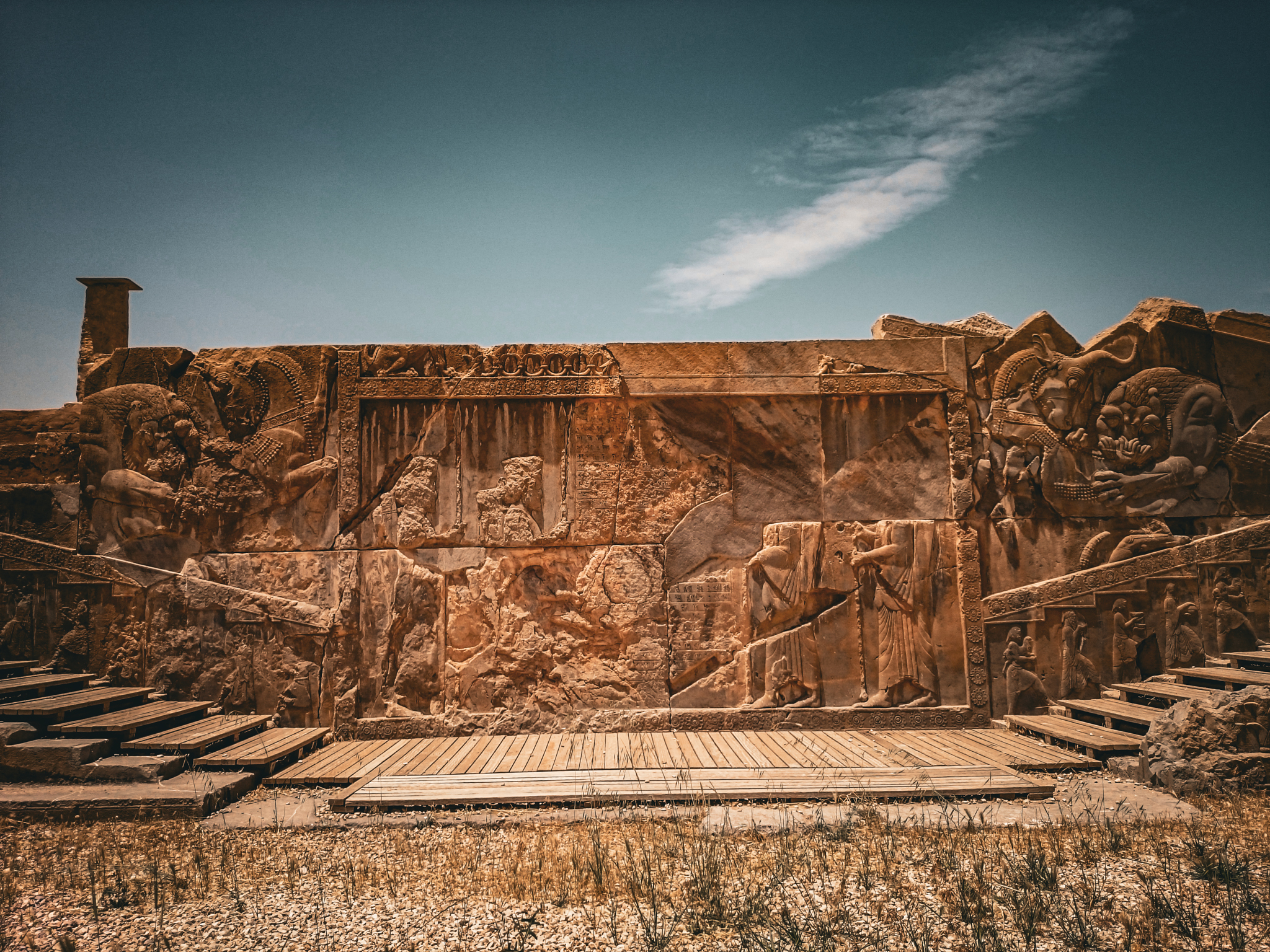 A close-up of an ancient stone relief on the central wall of a grand staircase, depicting four Achaemenid soldiers in profile. Symmetrical carvings of a lion attacking a bull are visible on the stair-fronts.