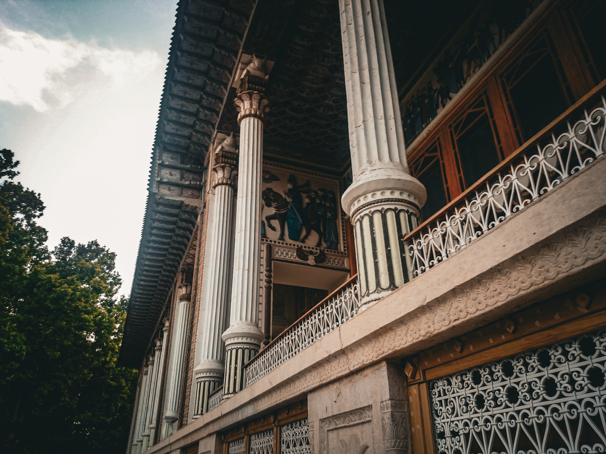 Low-angle outdoor view of the Afif-Abad Garden porch in Shiraz, featuring four tall stone columns with carved capitals and a wooden ceiling with floral patterns.