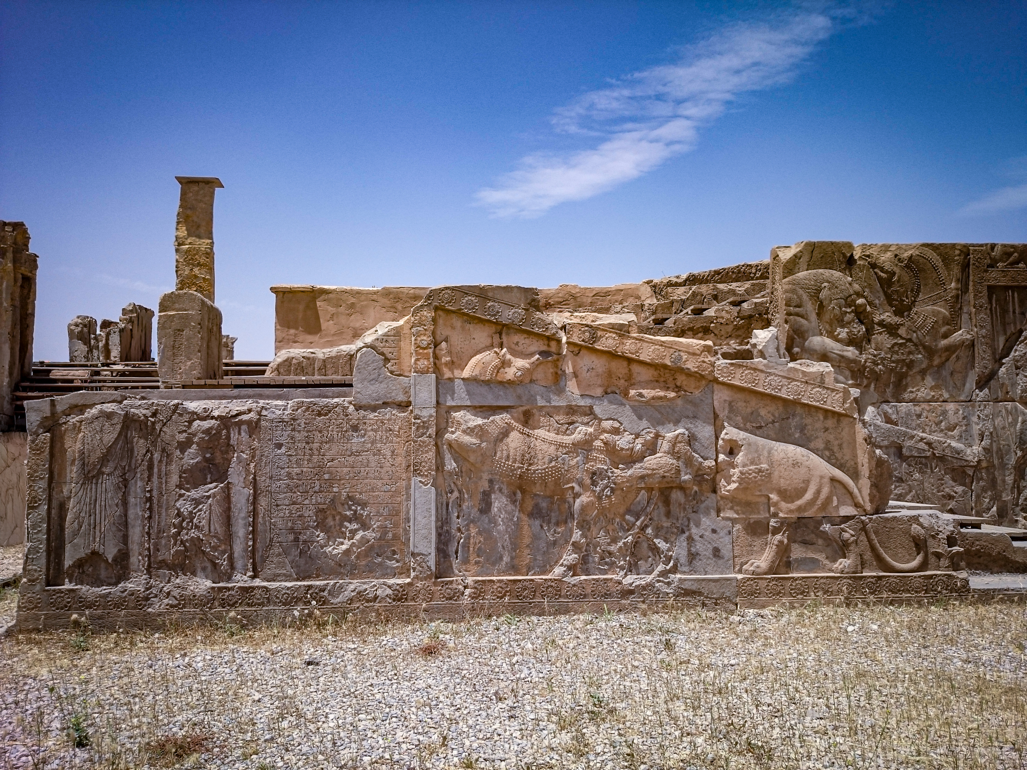 A stone bas-relief at Persepolis showing a lion attacking a bull, with a cuneiform inscription and two partial figures of soldiers on a staircase wall.