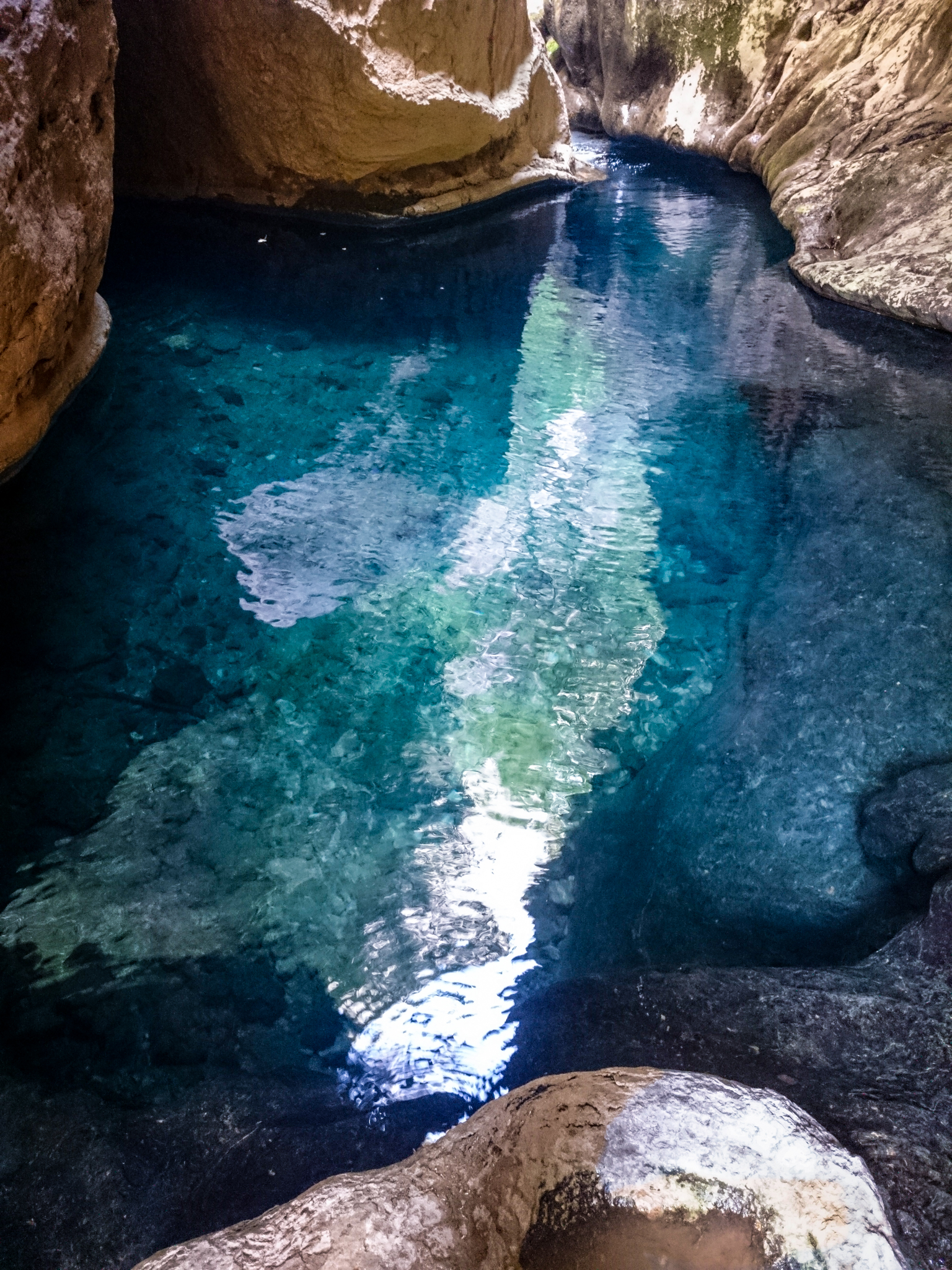 A small, clear blue lake is completely surrounded by mountains, reflecting the green foliage and the cream-colored base of the slopes in its calm water.